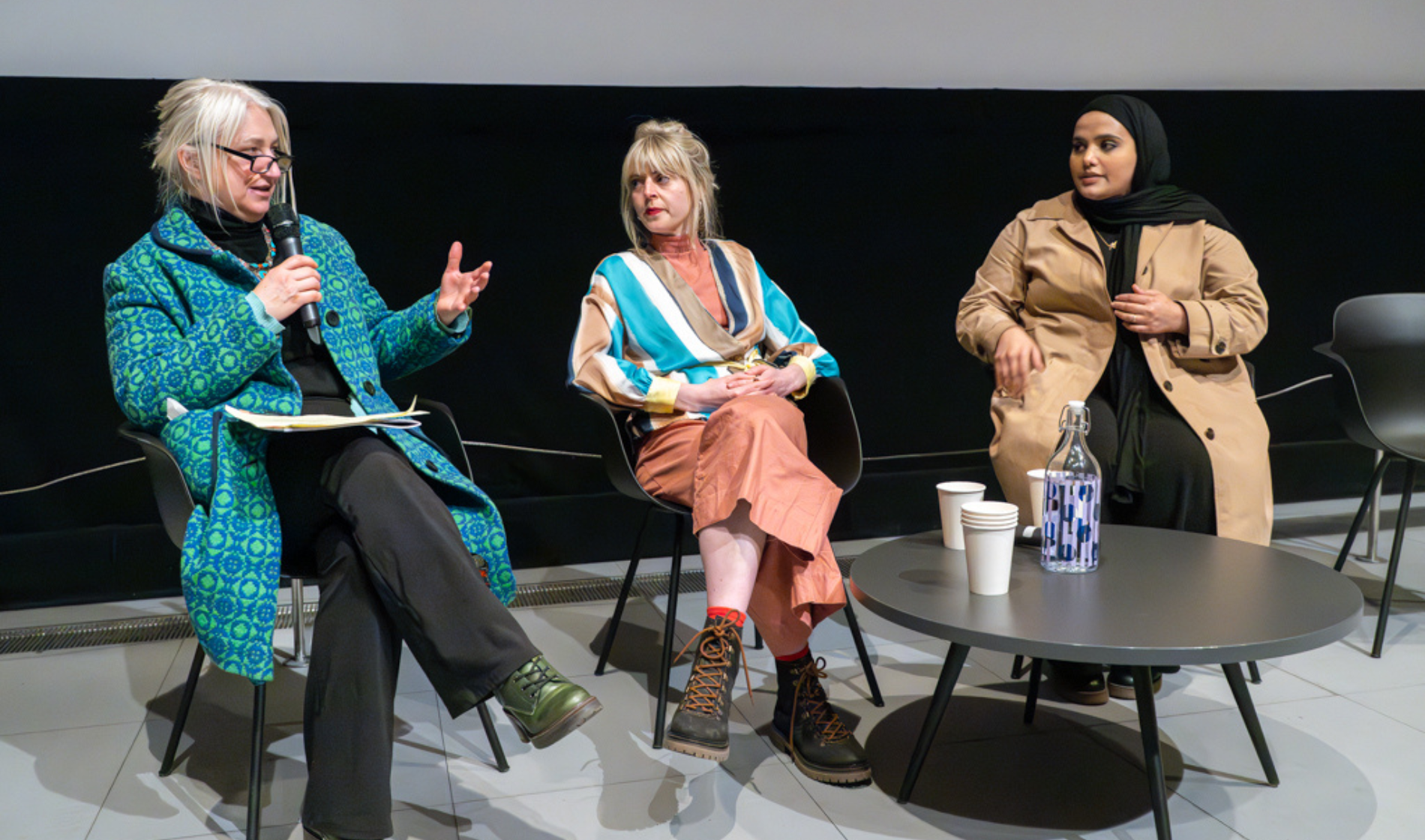 Zoe Partington, Dora and Amina sit with a screen behind them and a mic in panel format