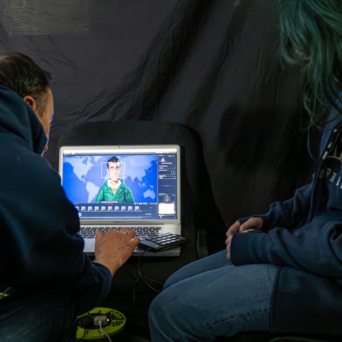 A member of Twin Vision is seated with a young person looking at an animated man on screen looking like he is ready to read the news