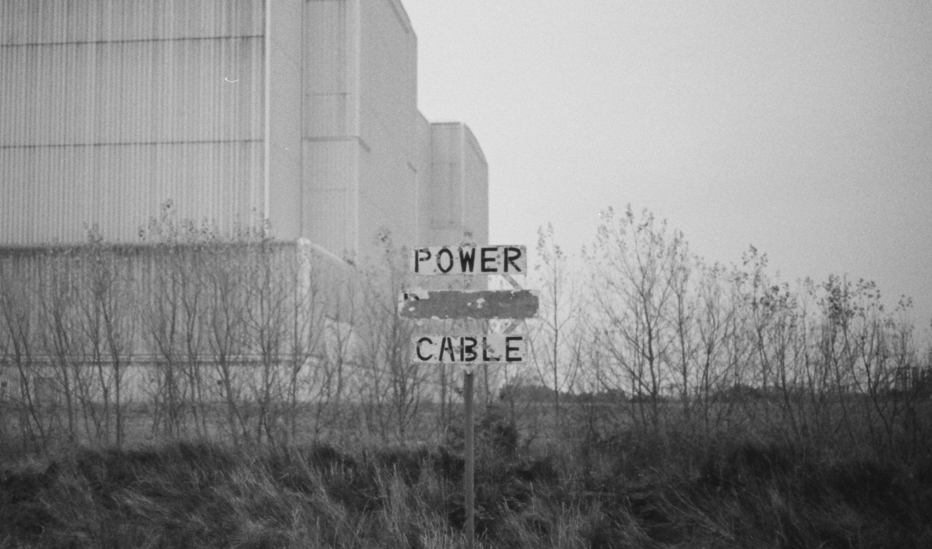 A sign standing in front of some kind of desolate factory building reads 'Power (scribble) cable)