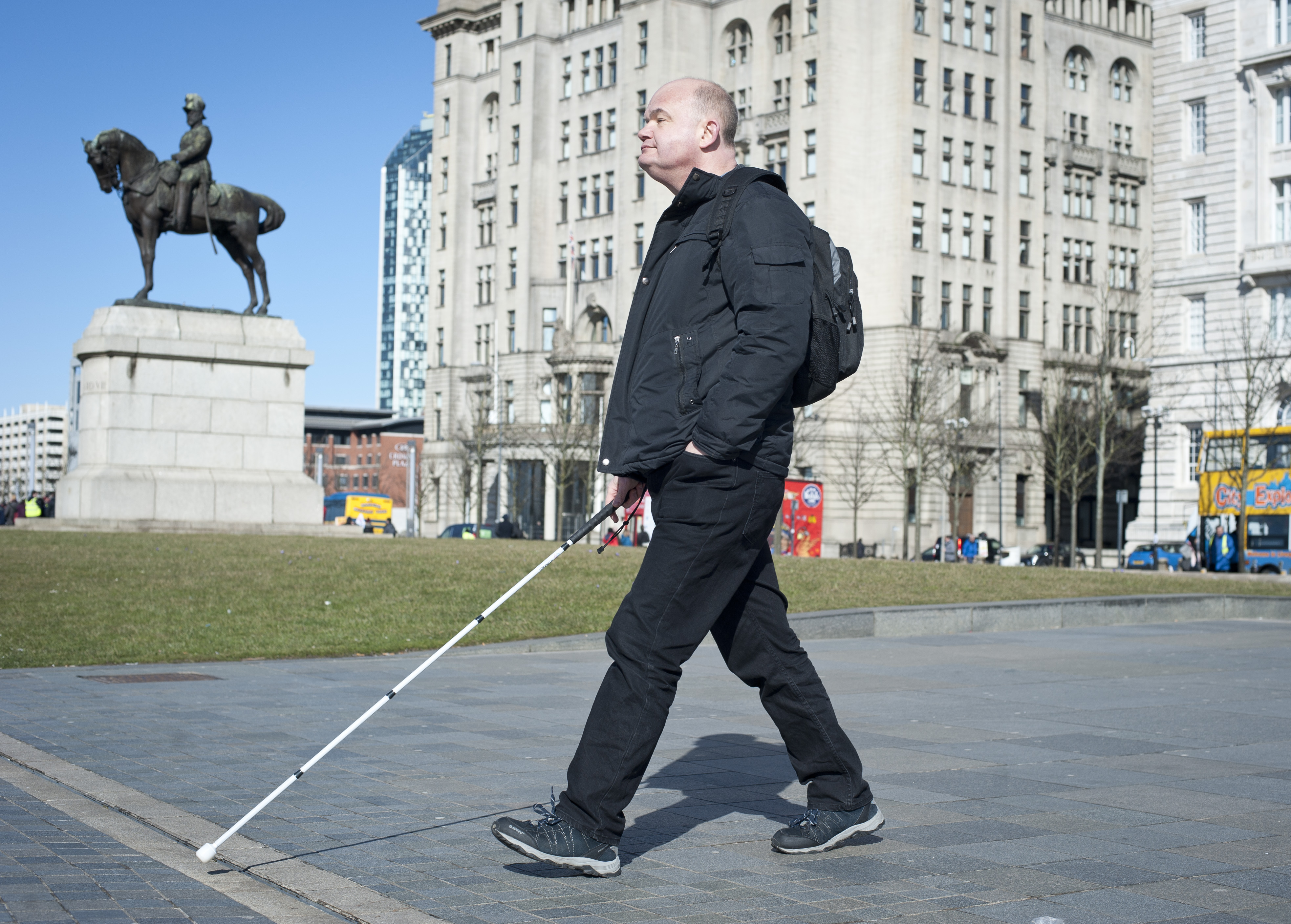 Tom Walker walks in front of the three graces with his white cane on a sunny day with blue sky.